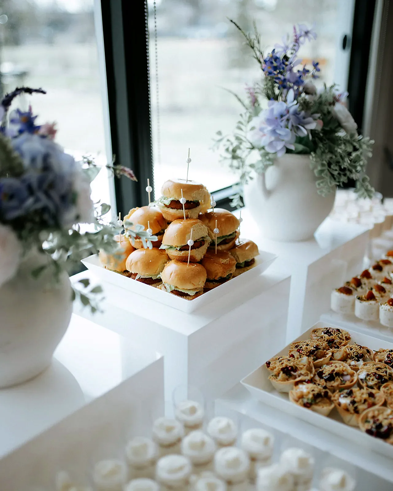 Sliders, dessert cups and florals on white risers by a window, a styled catering display by Deep's Delights